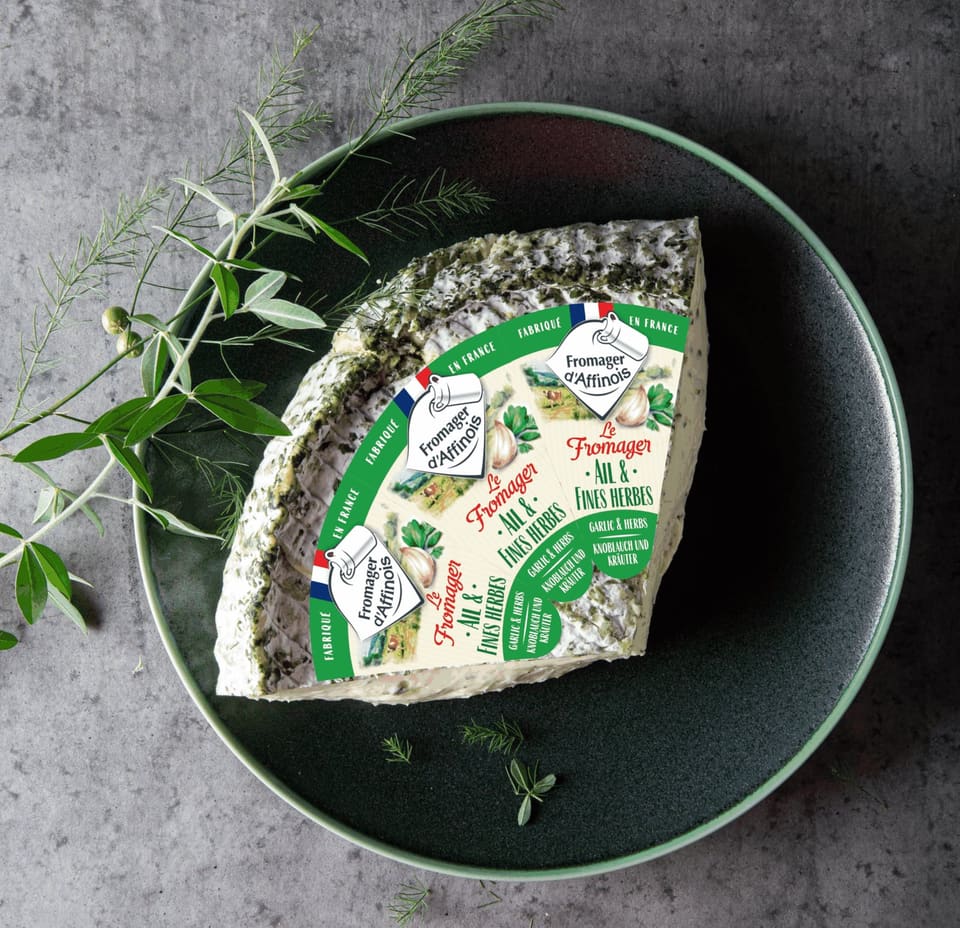 Клин мягкого сыра Fromager d’Affinois в фирменной обёртке на тёмной тарелке с травами