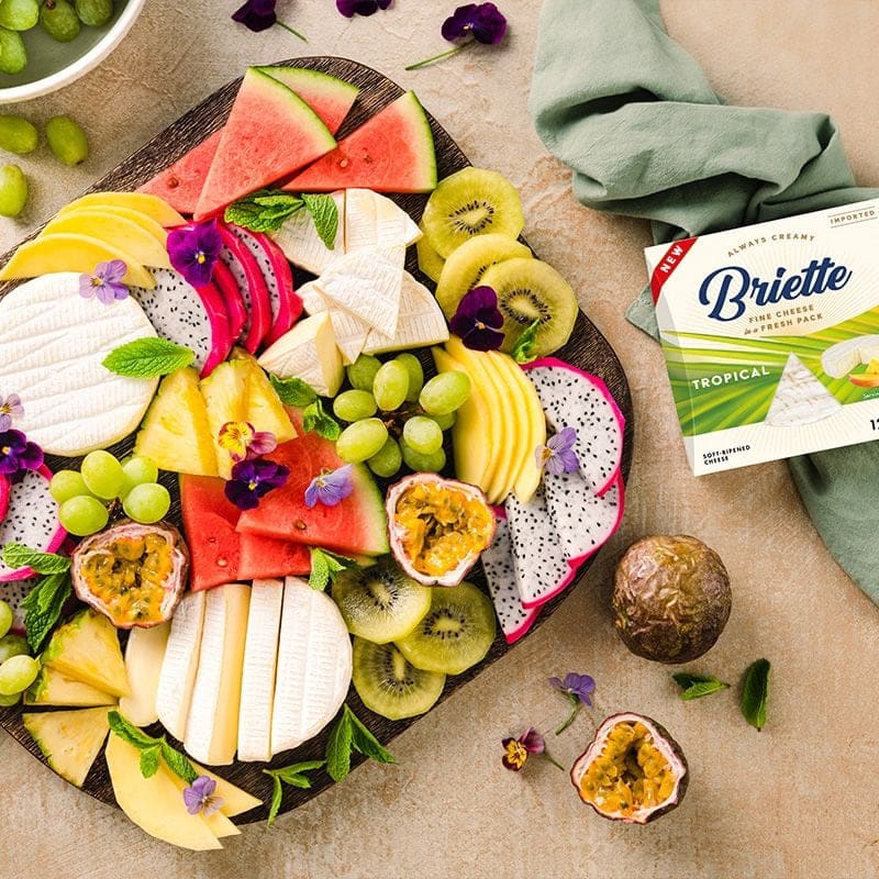 Доска с нарезанными фруктами и ломтиками мягкого сыра рядом с коробкой Briette Tropical