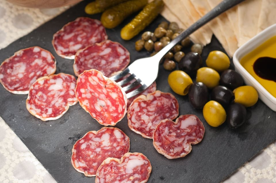 Ломтики сальчичона на тёмной доске с оливками, корнишонами и маслом