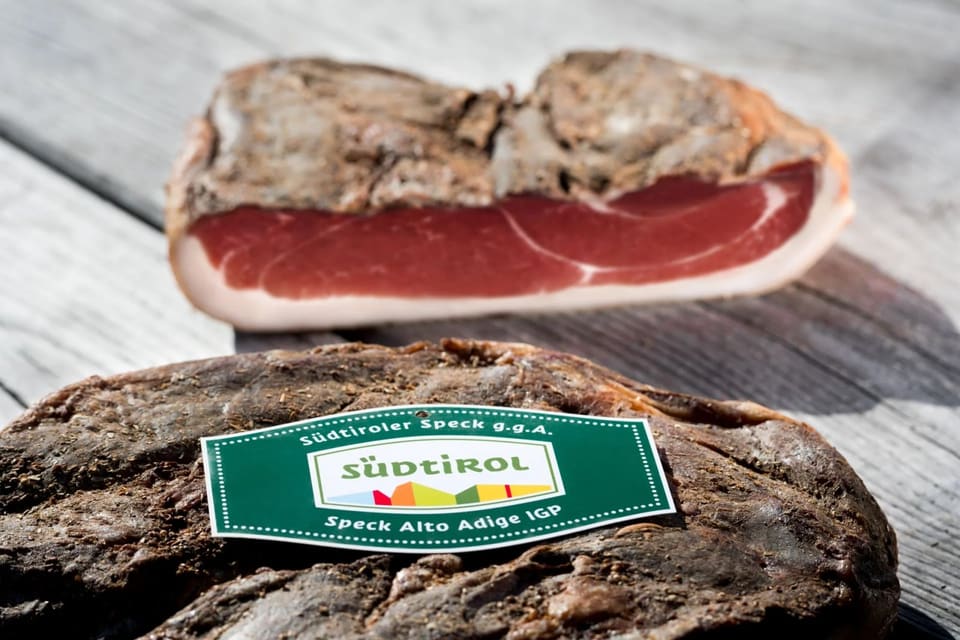 Кусок шпека на корке, рядом этикетка Südtiroler Speck g.g.A. / Speck Alto Adige IGP